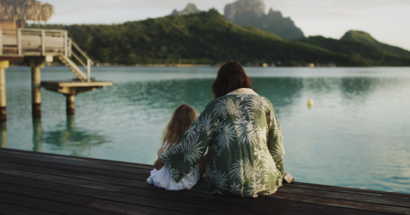 Brunette man sitting down with blonde child overlooking sea with big mountains behind