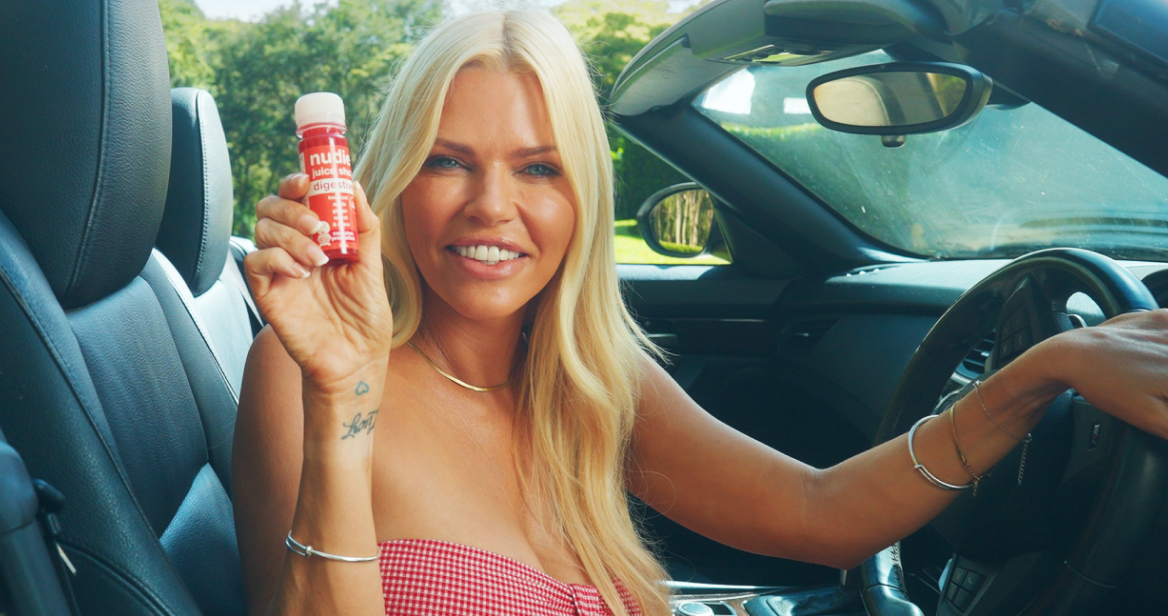 Sophie Monk smiling while holding a nudie 60ml juice shot in a convertible car, as part of Circul8’s cheeky campaign for nudie juice