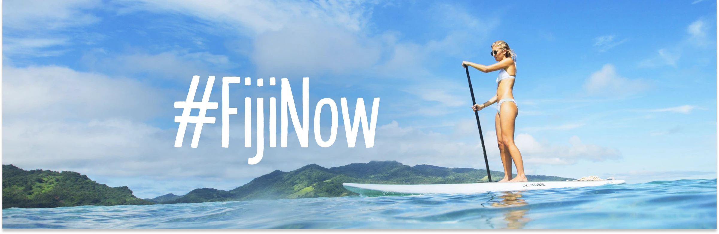 fijinow-campaign_Tourism-Fiji_circul8-work-long-1193-384 A woman in a white bikini and sunglasses stands on a paddle board in the ocean, holding a black paddle. The hashtag #FijiNow is written in large, white, hand-drawn text on the left side of the frame. In the background, lush green tropical mountains rise under a bright blue sky with scattered white clouds.