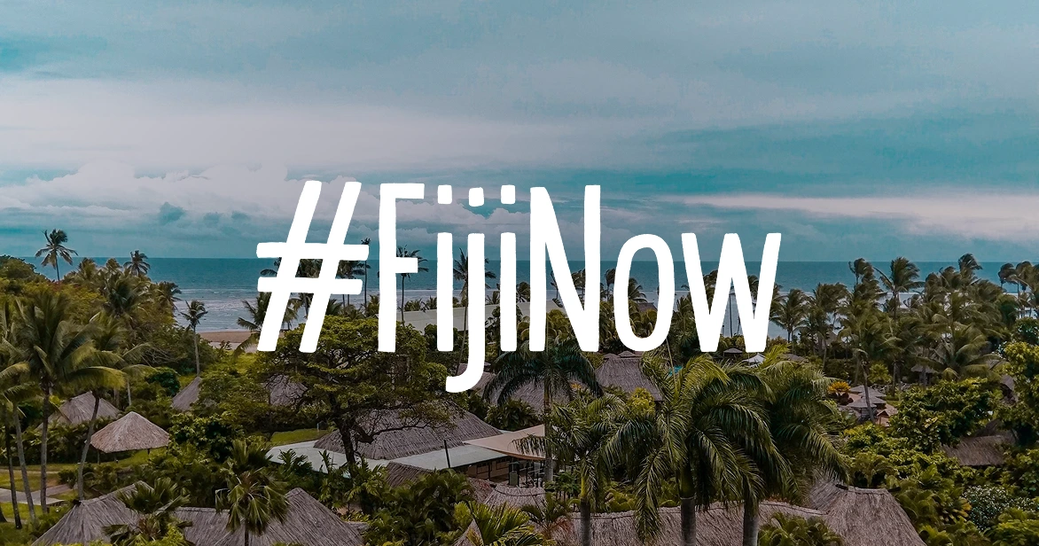 tourism-fijinow-bula-as-usual An aerial birds-eye view of a small, secluded tropical island in the middle of a deep blue ocean. The island is ringed by a white sand beach and a bright turquoise coral reef, with several overwater bungalows extending into the water.