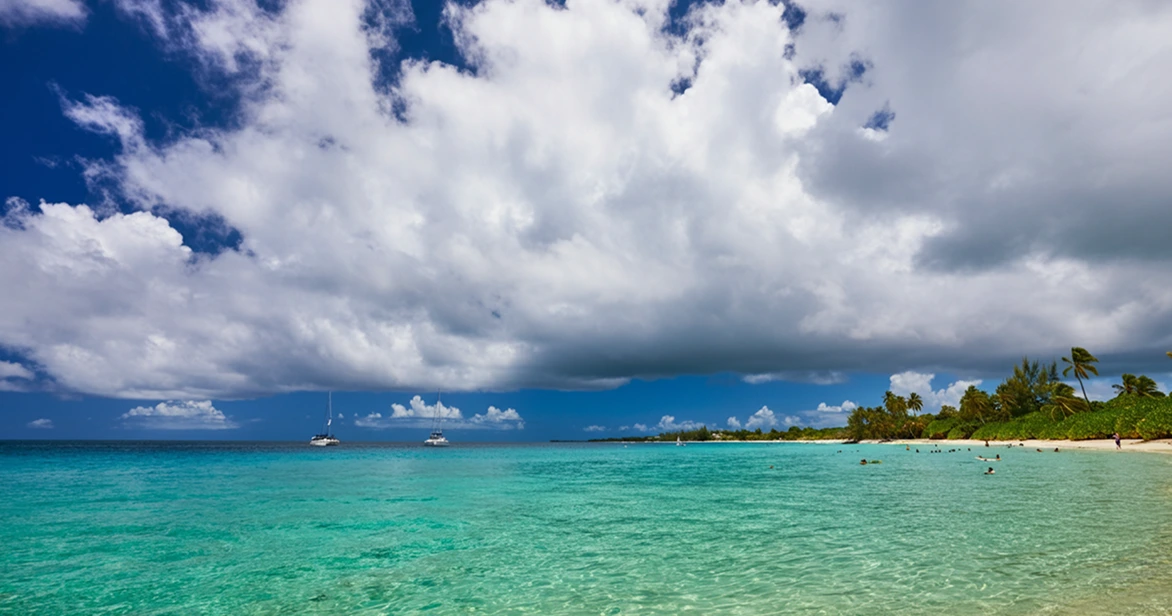tourism-fijinow-fiji-beaches cloudy sky A wide-angle shot of a beautiful Fijian beach with vibrant turquoise water. Large, dramatic white and grey clouds fill the sky over the ocean, where two white catamarans are anchored in the distance.