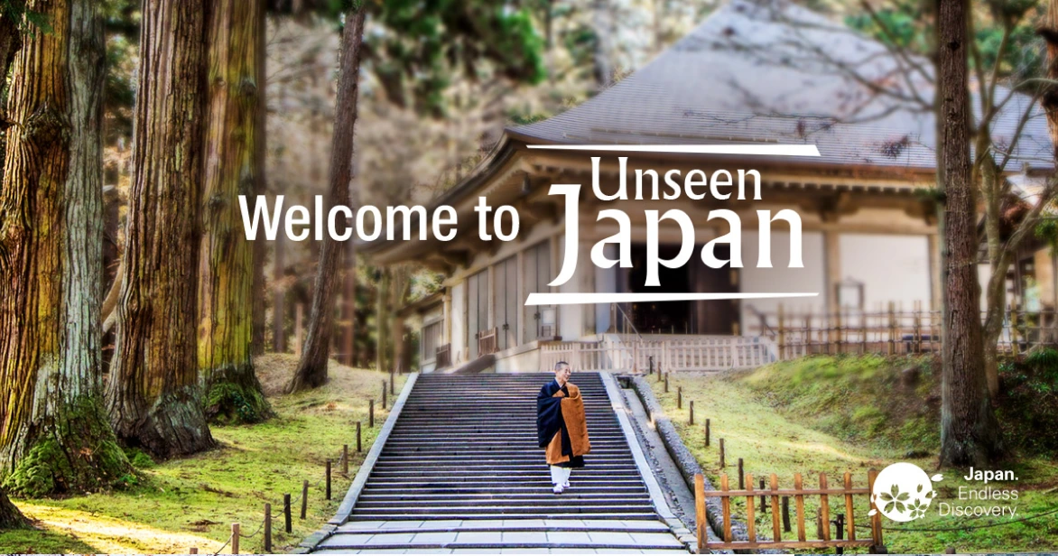 A Buddhist monk walking down stone steps of a traditional temple surrounded by tall cedar trees, with the "Welcome to Unseen Japan" logo and JNTO branding.