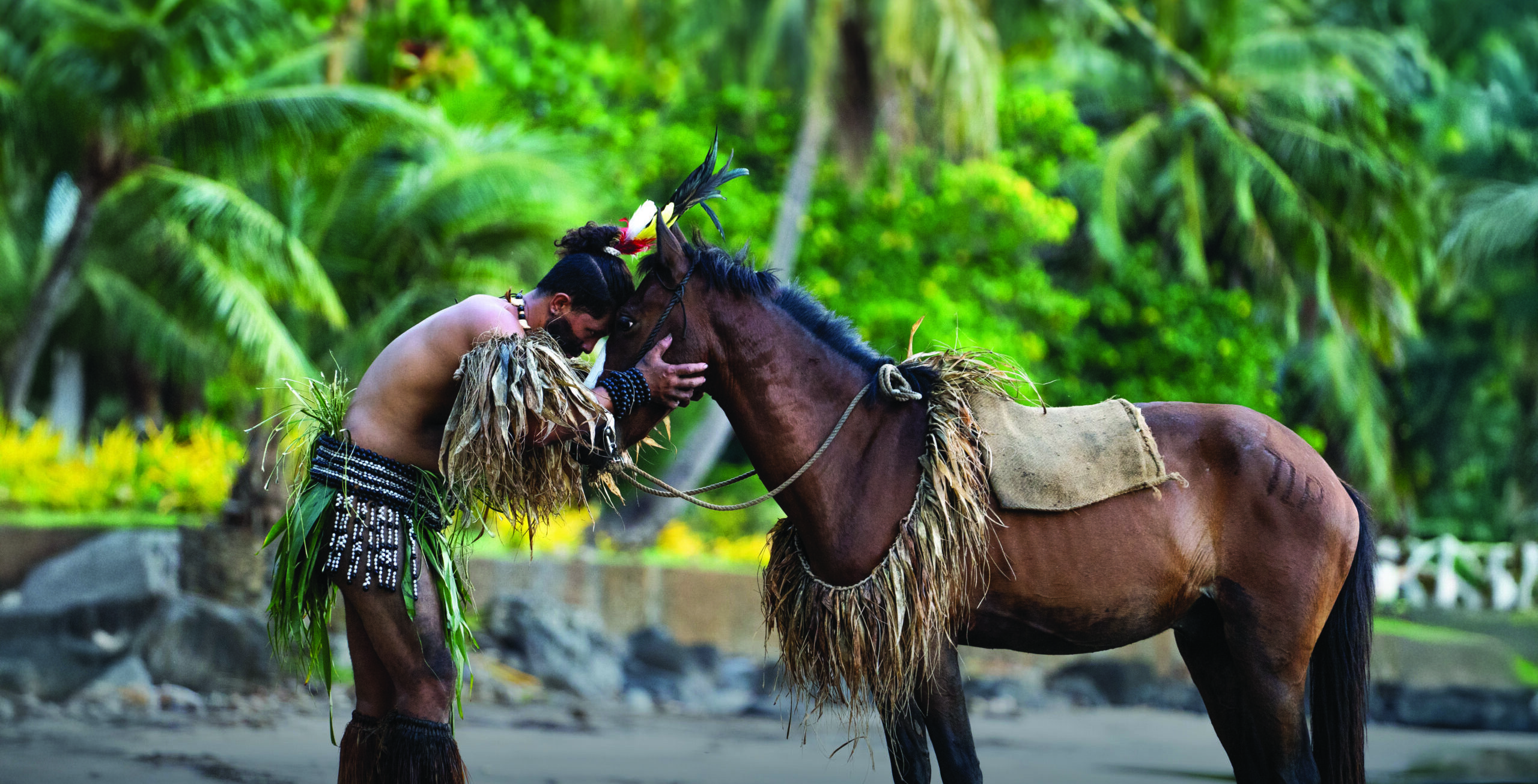 A Tahitian man in traditional attire gently touches foreheads with a horse on a tropical beach surrounded by palm trees.