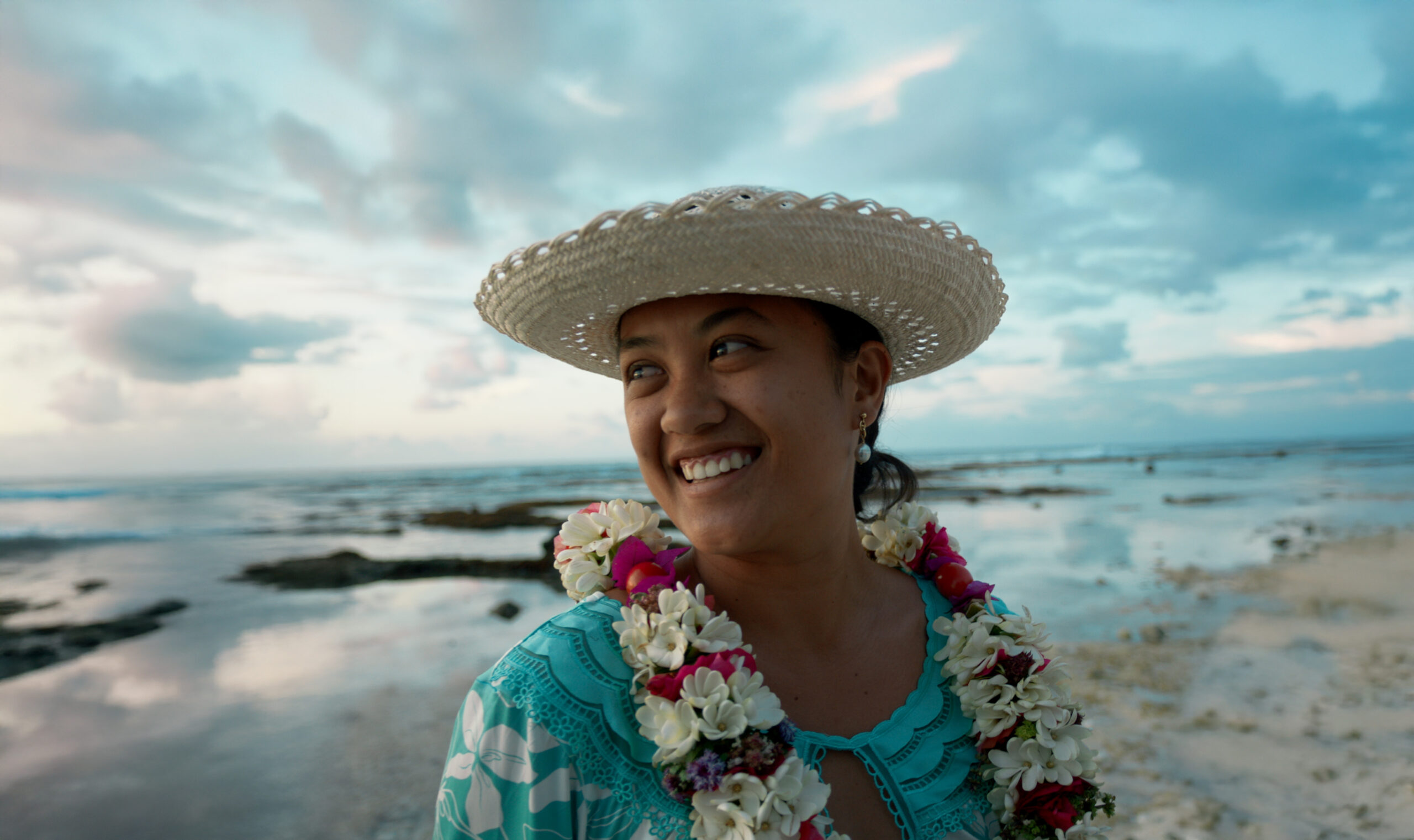A smiling Tahitian woman wearing a straw hat and flower lei stands by the ocean at sunset.