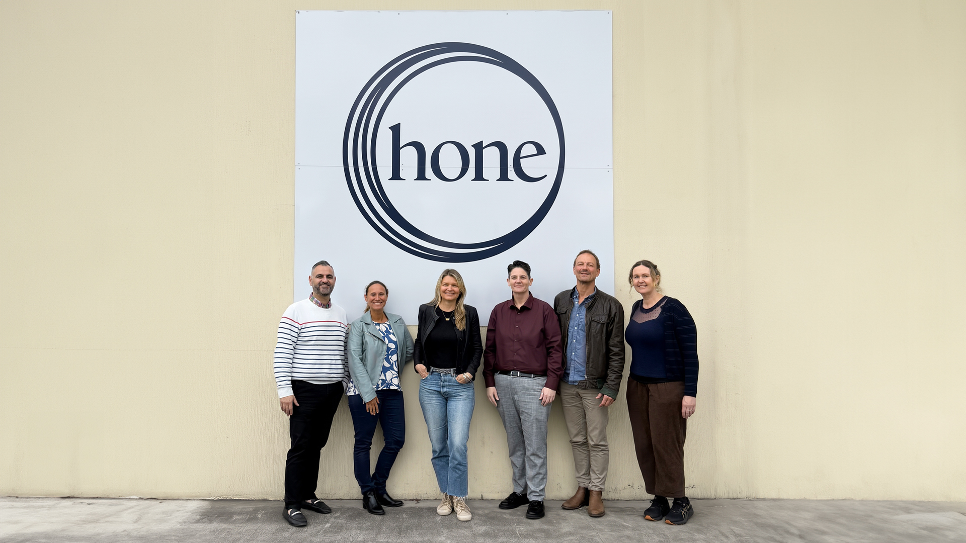 George Boutros (Chief Operating Officer, Hone), Audrey Wigsten (Head of Operations & Delivery, Circul8), Alana Stocks (Managing Director, Circul8), Nicole Bruce (CEO, Hone), Phil Watson (Creative Strategy Partner, Circul8) and Julie Floyd (Head of Production, Circul8) standing in front of a large Hone logo sign, celebrating their new creative and marketing partnership.