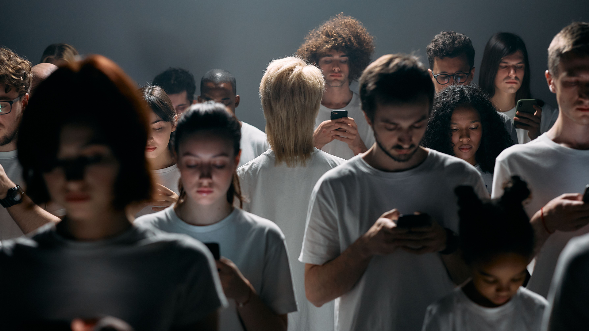 A crowd of people in white t-shirts standing in a dim, smoky room, each looking down at their phone