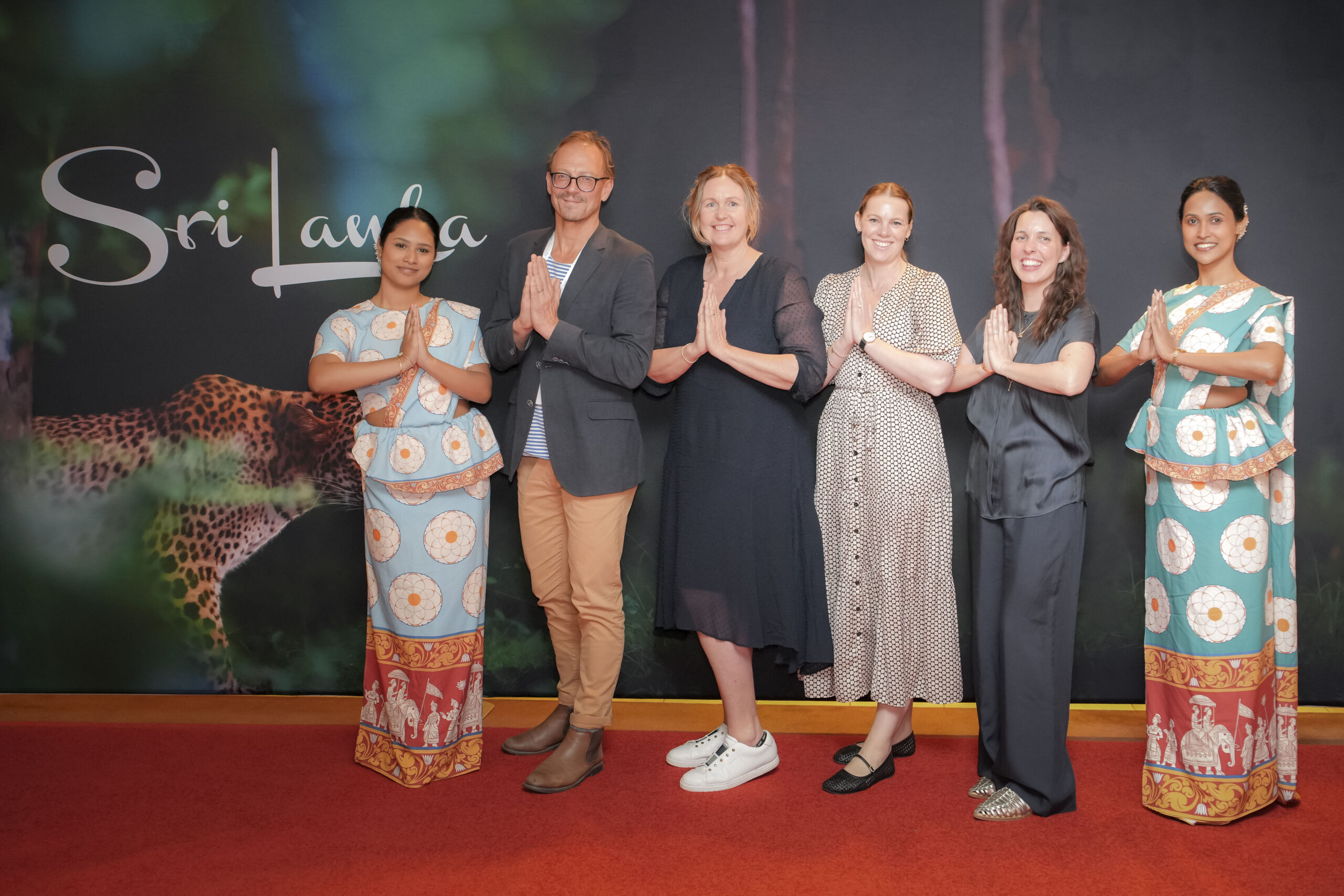 Phil Watson, Julie Floyd, Abbey Sands and Jessica Cory greet with hands in namaste in front of a Sri Lanka backdrop featuring a leopard, two in traditional Sri Lankan dress